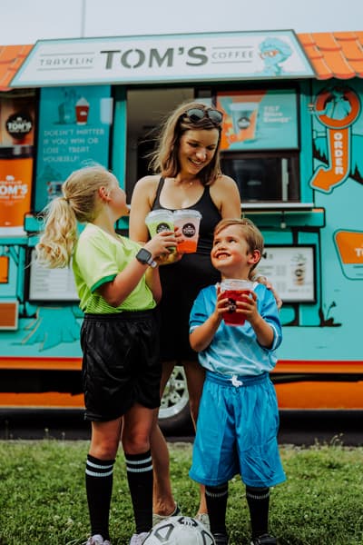A mother and two kids in soccer uniforms holding Travelin' Tom's cups at the truck