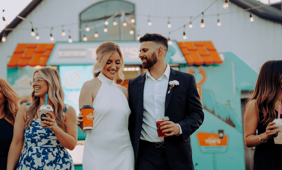 A bride and groom holding branded Travelin' Tom's coffee cups under café lights at their wedding